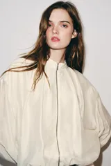 Chaqueta bomber de cuello subido y manga larga, con acabado satinado en color beige. Presenta pliegues frontales, bolsillos laterales ocultos en costura y cierres elásticos en puños y cintura. Cierre frontal con cremallera.