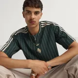 Camiseta de punto oversized verde oscuro de la colección Icons de Adidas, inspirada en los años 80, con rayas diplomáticas blancas, las icónicas tres tiras de la marca en las mangas, cuello redondo y el logotipo del trébol bordado junto al escudo vintage de la Juventus.