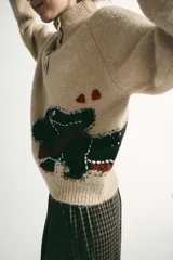 Jersey de punto color beige con cuello subido y cierre frontal de botones. Presenta un bordado de un perro salchicha con una bufanda roja y corazones en la parte inferior delantera.