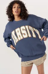 Sweater de tejido de punto color azul oscuro jaspeado, con cuello redondo y hombros caídos. Presenta la palabra "VARSITY" estampada en el frente con tipografía de bloque en color crema y borde naranja.