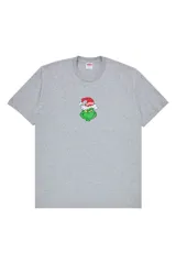Remera de algodón gris jaspeado con cuello redondo y manga corta. Presenta un estampado central pequeño que muestra al personaje Grinch usando un gorro de Navidad con el logo de Supreme.