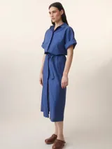 Vestido camisero midi azul con rayas verticales, cuello camisero, mangas cortas con puños y cinturón de lazo. Tiene dos bolsillos de parche en el pecho y cierre frontal con botones.