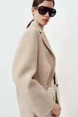 Abrigo corto de Zara confeccionado en hilatura con lana, de color beige. Presenta cuello solapa, manga larga, cierre frontal con botones negros y bolsillos de plastrón en el delantero.