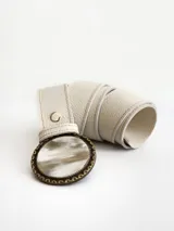 Cinto de cuero color crudo con forro de cuero y sistema de broche suizo intercambiable. Presenta una hebilla circular de guampa con borde metálico labrado en tono bronce.