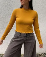 Remera de morley de algodón color mostaza, de manga larga y ajuste al cuerpo. Presenta cuello alto y terminaciones con borde fruncido en cuello, mangas y ruedo.