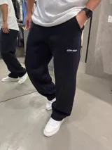Pantalón deportivo de corte baggy y estilo oversize, color negro, confeccionado en algodón. Presenta una costura vertical frontal y un pequeño logo bordado en la parte superior de la pierna izquierda.