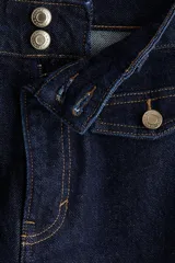 Jean de denim de algodón con leve stretch, color azul oscuro (índigo). Presenta un corte ajustado en la cintura y cadera, ensanchándose en un corte acampanado desde la rodilla hasta el bajo. Es de tiro alto con cierre y botones frontales, y cuenta con solapas decorativas en los bolsillos delanteros y bolsillos parche traseros.