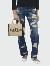 Cartera tipo tote bag de lona color beige, con diseño estructurado y asas superiores. Presenta el logo de la marca estampado en la parte frontal y detalles de cintas laterales con inscripciones.