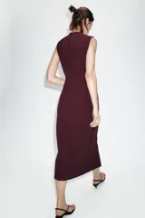 Vestido midi de cuello redondo y sin mangas, confeccionado en tejido tipo crepe. Presenta cierre frontal con botones dorados, bolsillos con solapa en la parte delantera y un cinturón elástico integrado con hebilla metálica decorativa.