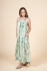 Vestido largo de corte holgado con escote halter y lazo ajustable en el cuello. Presenta un estampado tipo tie-dye en tonos verdes y crema, con un cinturón fino del mismo tejido para ajustar la cintura y bolsillos laterales.