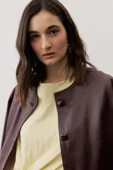 Chaqueta de cuero negra con cuello redondo, cierre frontal de botones forrados y bolsillos ribeteados con detalles de costura decorativa.