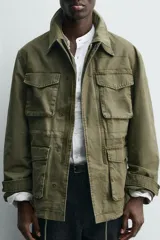 Cazadora de estilo militar en color verde oliva, confeccionada en algodón con efecto lavado. Presenta un diseño de corte relajado con cuello doble, cierre frontal con cremallera oculta por solapa con botones a presión, cuatro bolsillos frontales de plastrón con solapa y puños ajustables con botón.