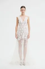 Vestido largo de fiesta con escote en V, confeccionado en tul transparente con aplicaciones de encaje floral blanco. Presenta un diseño sin mangas con detalle de volado en uno de los hombros y falda con caída fluida.