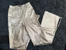 Pantalón de tiro alto confeccionado en eco cuero con acabado metalizado dorado. Presenta un corte recto con aberturas laterales con cierre en los tobillos y costuras frontales decorativas.