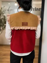 Chaleco de pana con diseño color block en tonos mostaza y bordó. Presenta un volante estampado en los hombros y en la parte superior de la espalda, con una etiqueta de cuero sintético en el centro.