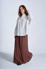Camisa blanca de textura arrugada con cuello alto con volados y lazo de ajuste, cierre frontal con botones de nácar, tablas decorativas en los hombros y mangas largas con puños elásticos.