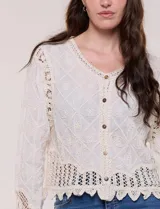 Cardigan corto beige de algodón con bordado en relieve, escote redondo y cierre frontal con botones carey. Mangas largas con detalles de encaje crochet y ruedo con terminación en ondas.