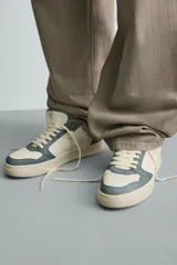 Zapatilla deportiva de estilo retro con diseño de paneles en combinación de colores crema, verde azulado y detalle en rojo en el talón. Presenta suela con volumen y cierre con cordones.