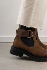 Bota tipo Chelsea de cuero marrón con suela track negra de goma. Presenta paneles elásticos laterales y una costura reforzada en la puntera. Incluye una correa de cuero desmontable con hebilla metálica.