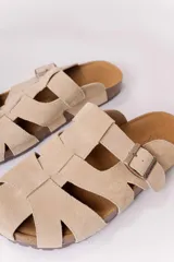 Zuecos de gamuza color beige, con diseño tipo sandalias romanas de tiras anchas entrelazadas, hebilla lateral metálica regulable y suela anatómica de corcho.