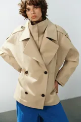 Trench corto oversize color beige con cuello subido de efecto doble, cierre cruzado con botones oscuros y bolsillos de vivo delanteros.