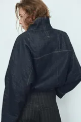 Cazadora de denim color azul oscuro, con cuello solapa, mangas largas tipo murciélago y bolsillos de vivo en el delantero. Presenta un bajo elástico ajustable con cordones y cierre frontal con cremallera y botones a presión ocultos.