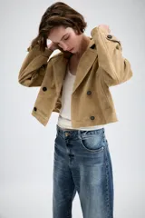 Trench corto cruzado de piel de ante color tostado, con cuello de solapa, bolsillos delanteros de vivo y manga larga con trabillas en los puños.