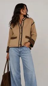 Chaqueta de lana merino color camel con detalles en contraste color marrón oscuro en los bordes, puños y bolsillos. Presenta cierre frontal con botones y cuello redondo.