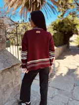 Sweater marrón de lana con cuello alto y manga larga. Presenta un copo de nieve blanco en el frente y bordados a mano. Tiene puños y cuello con diseño geométrico en tonos blanco, marrón y negro.