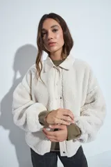 Campera de corderito color beige, con cuello camisero y cierre frontal de botones a presión. Presenta una cartera central lisa en contraste y mangas largas con puños elásticos.