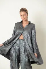 Camisa plateada de poliéster con textura metalizada, corte amplio, cuello clásico, cierre frontal con botones y mangas largas con puños abotonados.