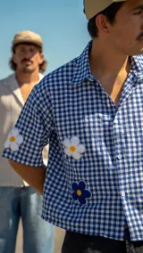 Camisa de corte boxy con estampado de cuadros vichy en azul y blanco. Presenta mangas cortas y aplicaciones bordadas de flores en relieve en color blanco y azul con centro amarillo.