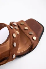 Sandalia de taco cuadrado medio, confeccionada en cuero vacuno negro. Presenta tiras cruzadas sobre el empeine decoradas con tachas metálicas doradas y cierre con hebilla ajustable en el tobillo.