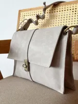 Bandolera de cuero color beige claro, con solapa frontal, cierre metálico de broche y correa regulable con detalles ondulados.