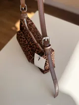 Cartera tipo baguette con estampado animal print, confeccionada en material suave símil gamuza. Presenta un bolsillo frontal con solapa y cierre, compartimento principal con cierre superior, y asa larga regulable de cuero sintético marrón.