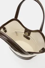 Bolso tipo bucket de diseño estructurado, confeccionado en combinación de canvas color crudo y cuero sintético marrón. Presenta doble asa de mano, pespuntes a contraste y base reforzada.