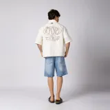Camisa de manga corta color marfil, corte boxy, con cuello camisero, cierre frontal de botones y pequeño bordado marrón en el pecho con las siglas VCP.