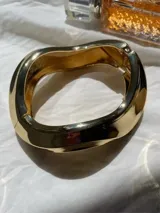 Brazalete rígido de diseño orgánico y asimétrico con acabado metálico brillante en tono dorado. Cuenta con un sistema de cierre de bisagra integrado.