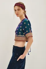Camisa de seda con diseño tipo chaqueta corta, de color azul petróleo con bordados decorativos en tonos dorados y rojos. Presenta mangas largas abullonadas de color crema y cierre frontal.