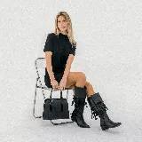 Bota texana de caña alta confeccionada en cuero vacuno negro. Presenta un diseño con flecos decorativos en la parte superior de la caña, taco cuadrado de madera y punta fina. Incluye cierre lateral interno.