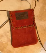 Mini bolso de gamuza color naranja rojizo, con solapa y cierre de cordón de cuero. Presenta un detalle de costura decorativa en los bordes y un parche de cuero marrón con el logo "Sierra Mora".