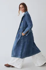Trench largo de denim azul, con diseño de doble botonadura, solapas clásicas y mangas largas con puños ajustables. Presenta un corte recto y estructurado.