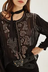 Camisa de manga larga en color negro con tejido semitransparente y detalles de hilos metalizados. Presenta un diseño con bordados florales en tonos tierra, cierre frontal con botones y un lazo decorativo en el cuello.
