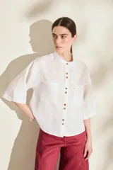 Camisa blanca de lino, con cuello mao, mangas 3/4 anchas, bolsillos frontales con solapa y cierre de botones marrones contrastantes.