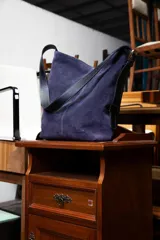 Bolso tipo bucket de gamuza color violeta con correa ajustable de cuero negro y detalles de costuras pespunteadas a la vista.