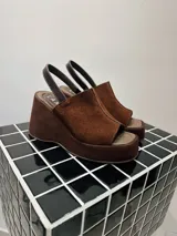 Sandalias de plataforma y taco ancho tipo wedge, confeccionadas en cuero gamuzado color marrón chocolate. Presentan diseño destalonado con tira trasera y capellada cerrada con apertura en la puntera (peep toe).
