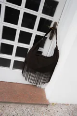 Bolso tipo hobo de gran tamaño en color marrón, confeccionado en gamuza. Presenta un diseño con flecos largos en la parte inferior, cierre superior y una correa trenzada con detalles artesanales.