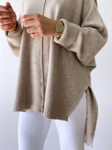 Buzo de tejido de punto color visón, con diseño holgado, cuello redondo, mangas largas con puños acanalados y costura central vertical visible. Presenta un ruedo asimétrico con aberturas laterales.