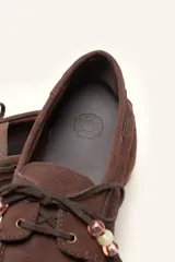 Mocasín estilo náutico de gamuza marrón, con cordones de cuero, borla decorativa y cuentas de colores en el empeine.