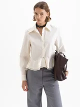 Camisa blanca de popelina con ajuste al cuerpo, puños anchos, cuello de puntas largas, broches dorados y doble volado en la cintura.